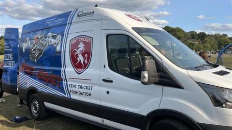 A minibus with a Kent Cricket badge on the side. It is half white and half blue. It has a games console on the side.