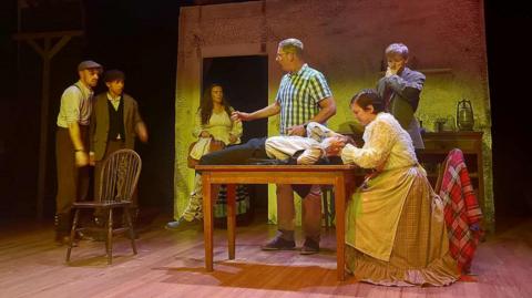 A scene from the play in which seven actors appear in a kitchen. They wear clothing from the 1830s - the men are in braces and flat caps while the women wear long skirts and aprons. One man lies on a table and holds his head, another looks on with a concerned expression.
