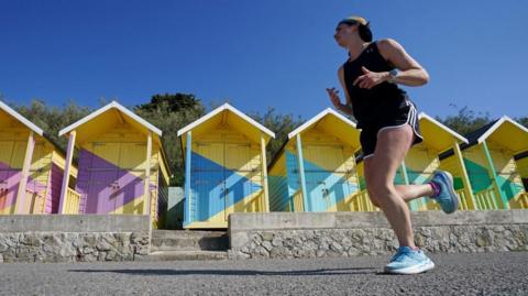 A runner enjoys the morning sunshine in Folkestone, Kent. Striking yellow beach huts with purple, blue or green arrow designs on their front are behind him.