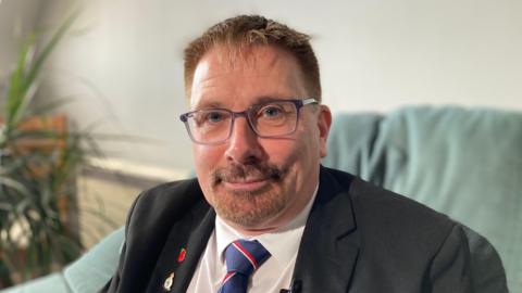 A 58-year-old man with short light brown hair and glasses wears a suit with a white shirt, naval tie and poppy. He sits on a green sofa with a pot plant just visible.