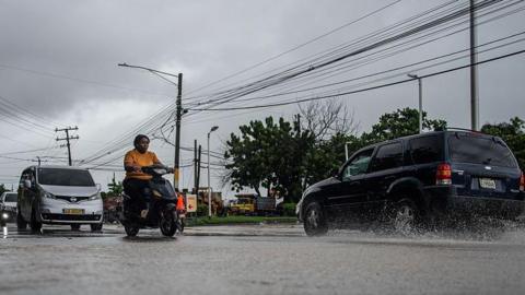 Cars and a motorbike drive on a street in the Dominican Republic amid rain. Power lines can be seen overhead 