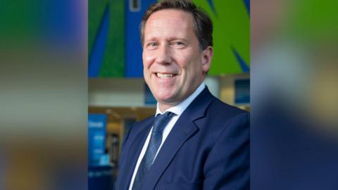A man with brown and grey hair wearing a white shirt and navy tie and suit jacket.