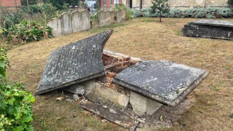 A tombstone which has been vandalised. The top has been smashed in two with one half sitting at an angle. There are gravestones in the background and another tombstone.