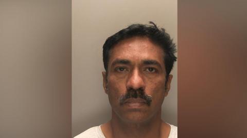 A police head and shoulders photo of a man with short black hair and a moustache, pulling a blank expression and wearing a white top against a grey wall.