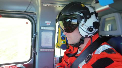 A white middle-aged man wearing a crash helmet and orange overalls, sitting in the rear of a helicopter, next to a window