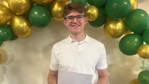 Leo Crowe wears a white shirt and dark spectacles. He holds a piece of paper with his results and stands under an arch with green and gold balloons.