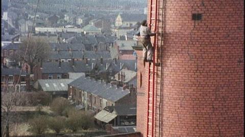 A wide shot of Fred Dibnah climbing a ladder up a very tall, red brick chimney.