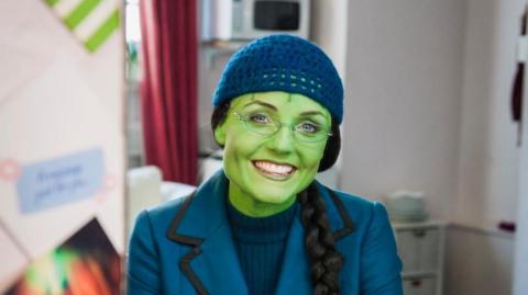 Kerry Ellis sits in a dress room in a chair and smiles at the camera. She has green face paint on to match her character Elphaba. She wears a blue blazer, blue jumper underneath and a blue hat. She is also wearing a black wig that has been braided. 