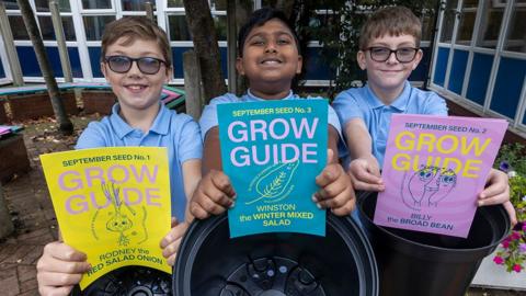 Three school boys in pale blue polo shirts smiling at the camera and holding brightly coloured leaflets that say "grow guide"