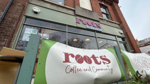 The exterior of a coffee shop called Roots. It is a brick, end of terrace building with olive green windows and a sign with pink writing on saying "Roots". There is a red, green and beige banner outside the café, bearing its name. The sky above it is wintry and cloudy.