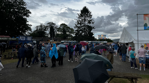 Crowds of people at a festival. Many are holding umbrellas or wearing raincoats. Food and drink stalls are on the left and right, with portaloos in the background