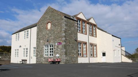 A school building with white walls and a section of open brickwork with a large playground in front of it and a blue sky with some cloud above