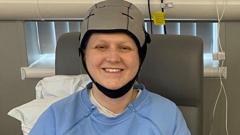 Close-up photograph of Lisa, who is smiling as she is sitting on a bed. She is wearing a grey scalp-cooling cap and a pale blue sweatshirt.