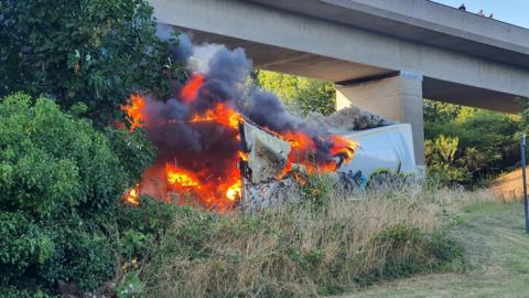 A lorry trailer on fire among greenery under a concrete bridge