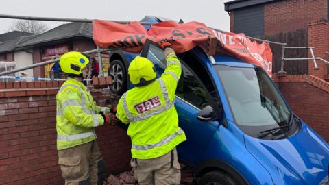 Two firefighters are pictured with a car hanging over a wall after a crash in a supermarket car park