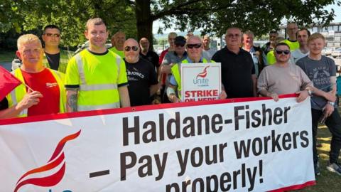 A group of men standing behind a white banner that says 'Haldane Fisher Pay Your Workers' in bold clack letters. 