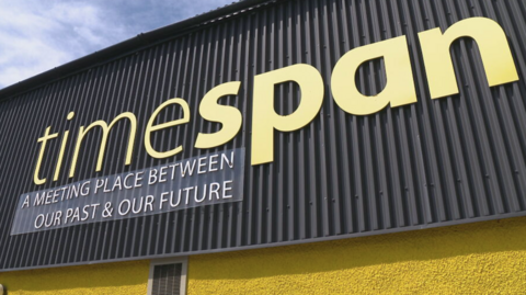 Part of the exterior of Timespan. There is an area of grey corrugated sheet above yellow painted wall. The corrugated sheet has the word "Timespan" in large yellow letter above a sign that reads: "A meeting place between our past and our future".