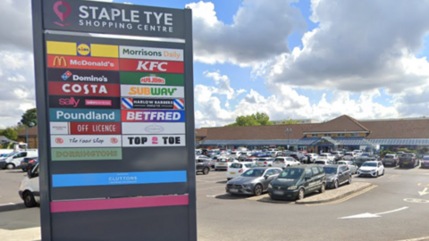 The car park at Staple Tye Shopping Centre in Harlow. On the left is a grey sign which reads "Staple Tye Shopping Centre", and the sign lists the shops and restaurants at the centre