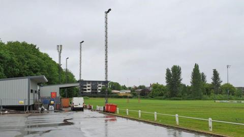 A rugby pitch with car park in the foreground. There is a block of flats in the distance, overlooking the club, and a club house and spectator stand to the left hand side of the image.