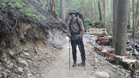 Sam Benastick in a undated police photo, seen on a dirt path in the woods wearing a backpack, hat and sunglasses and holding two hiking sticks