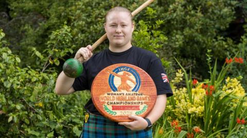Eve is wearing a green and blue kilt and holding a wooden disc with Woman's Amateur World Highland Games Championships 2025 engraved on it.