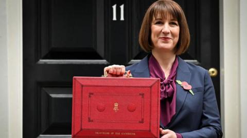 Chancellor Rachel Reeves stands outside Number 11, Downing Street.
She is dressed in a navy blue suit with a remembrance poppy in her lapel and a plum coloured blouse, holds a red box containing the Autumn Budget in October 2024. 