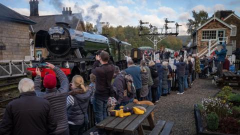 A crowd of people, including families and railway enthusiasts, gather on a station platform to admire and photograph the historic steam locomotive "The Flying Scotsman," numbered 60103. The engine emits a plume of steam as it prepares for departure. The station features traditional stone buildings, a wooden fence, and a signal gantry.