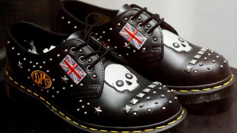 A pair of black shoes with yellow stitching, medal studs, an orange DMs logo in a circle, a small Union Jack flag and a skull incorporated into the design