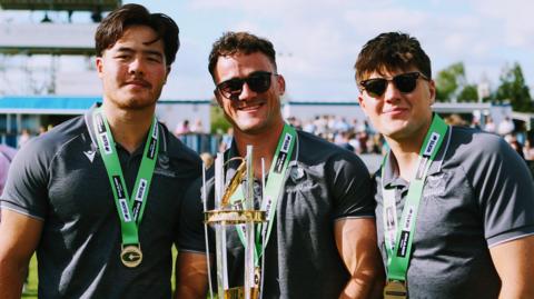 Three Worcester Warriors players celebrate winning the Champ Rugby title with the middle player Will Trewin holding the trophy