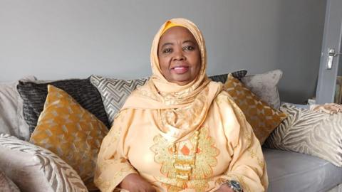 A woman sitting on a sofa wearing a headscarf and an embroidered gown.