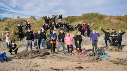 A group of about 30 children stand on the edge of a beach with dunes behind them.  They have been collecting rubbish and have netting at their feet and two are holding up a tyre. 