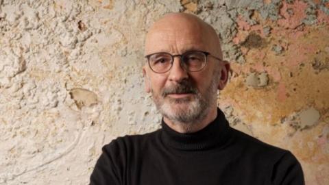 Trevor Avery is a bald man in his fifties wearing glasses and has a beard and moustache. He is standing in front of a wall that is heavily pockmarked with peeling paint that is coloured cream, pale pink and brown where it appears to be damp. He is wearing a black, roll-neck jumper. 