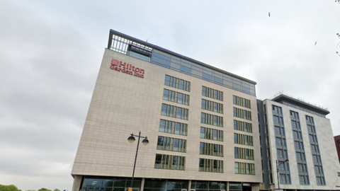 A roadside view of a tall beige building with glass windows and a red "Hilton Garden Inn" sign at the top.