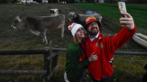 Dau berson yn sefyll mewn cae o flaen ffens gyda 3 buwch tu ôl iddyn nhw. Mae'r ddau gyda braich rownd ei gilydd wrth iddyn nhw gymryd llun. Ar y dde mae yna ddyn sy'n dal ei ffon lan gyda'i fraich chwith i gymryd y llun. Mae'n gwisgo het goch, felyn a gwyrdd gyda bathodyn tîm pêl-droed Cymru ar y blaen. Mae'n gwisgo crys-t'r tîm pêl-droed sy'n goch a siaced y tîm pêl-droed sydd eto'n goch. I'w dde mae yna fenyw sy'n gwisgo het wen gyda chalonnau coch arni. Mae hi hefyd yn gwisgo crys-t coch tîm pêl-droed Cymru a siaced wyrdd.