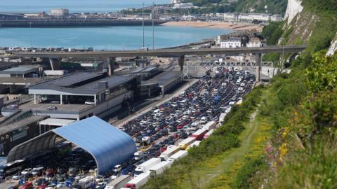Long, stationary queues of traffic on the approach to the Port of Dover, as seen from above. The town and its beach can be seen in the background on a sunny day.