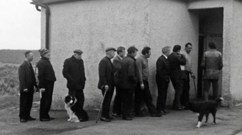 Black and white image of a line of men queuing outside a rural public toilet.