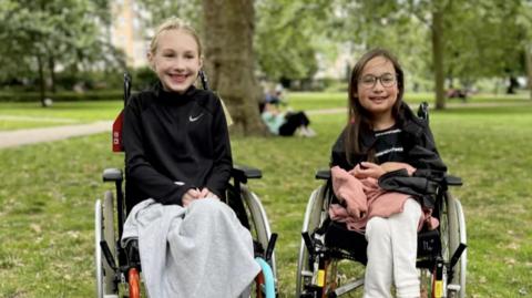 Two young girls one, with blonde hair and one with dark hair, sitting in wheelchairs in a park, smiling at the camera.