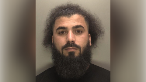 A mugshot of a man with a long fluffy beard and dark hair. He wears a black t-shirt and looks at the camera.