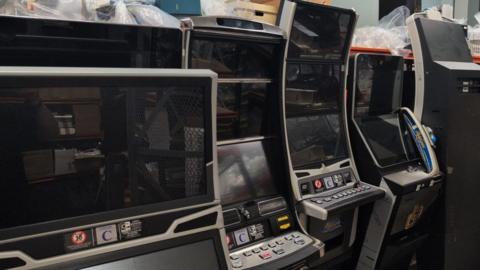A row of electronic gambling machines stored closely together, most with large black screens and control panels featuring rows of buttons. In the background are shelves and stacked items, including cardboard boxes and plastic-wrapped materials.