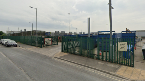 Hazel Court Household Waste Recycling Centre (HWRC)  in York, set back from a paved road, surrounded by green iron gates.