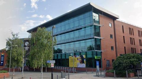 A google street view of a pedestrian area in front of a modern university building from the pavement. A four story red brick, glass fronted structure with a sign saying it is the Harrison Library and a street sign pointing to St Peter's parking. To the left is a sign for the sports centre. Tall trees reach several stories high and there are smaller ones growing behind a wall to the right.