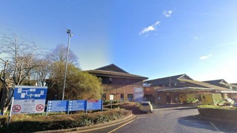 The Princess of Wales hospital on a sunny day as seen from the roundabout at the entrance. 
The brown brick two-storey building has a brown roof and canopy over the A&E entrance