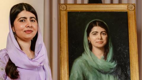 Malala Yousafzai is wearing a lilac dress and headscarf and is standing next to the new painting of her.
