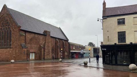 A road junction, to the left is a church building with shops next to it, on the right is a building with a black frontage and a sign that says kong