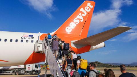 People boarding an EasyJet flight some carrying hand luggage and rucksacks on airport tarmac in Sardinia. It's a sunny day and the sky is blue.