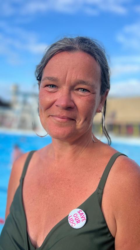 A woman in a green swimsuit with a 'save our lido' badge on it. She is smiling while standing in front of a pool.