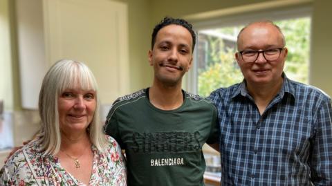 Anita is on the left of the picture. She has shoulder length white hair and is wearing a flowery blouse and a gold necklace. Aziz is in the middle and has short black hair and a green T-shirt that says "Sinners: Baleciaga". Steve is on the right of the picture and he is wearing a checked blue and dark blue buttoned shirt and he has dark glasses. He is balding but has a bit of short hair on his head.