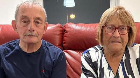 An elderly couple sit on a red leather sofa in a living room. The man on the left has cropped grey hair and a grey moustache, and wears a navy blue t-shirt and dark jeans. The woman has blonde bobbed hair, and wears red-rimmed glasses and a patterned blouse.