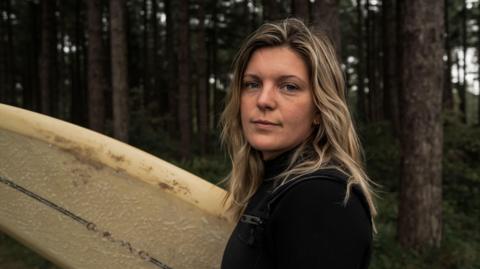 Laura Trulove in a wetsuit carrying her surfboard, she is looking at the camera and smiling. She has long blonde hair that looks dyed by the salt water and sun. The background is a forest.