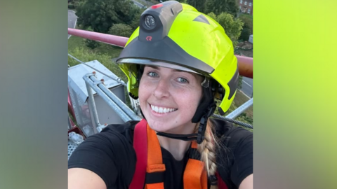 A smiling Jennie Logan's selfie as she is up a firefighting crane. Houses can be seen on the ground behind her. She is wearing a bright green helmet.